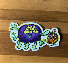 Load image into Gallery viewer, Colorful monster sticker with a basket of candy on a wooden surface. INKSHIFT.